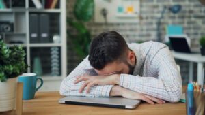 Man resting his head on a desk looking exhausted representing why am I always tired mental health and ongoing fatigue.